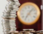 A close-up image of stacked coins with a blurred clock, symbolizing time and money relationship.
